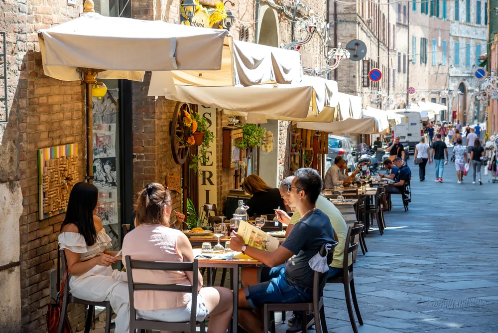 People dining outdoors at side on the street covered by an extended canopy from the restaurant, an images for an article that answers the question " is Tuscany safe to visit?"