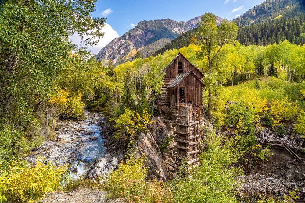 A cabin beside a river surrounded by forests and mountains. 