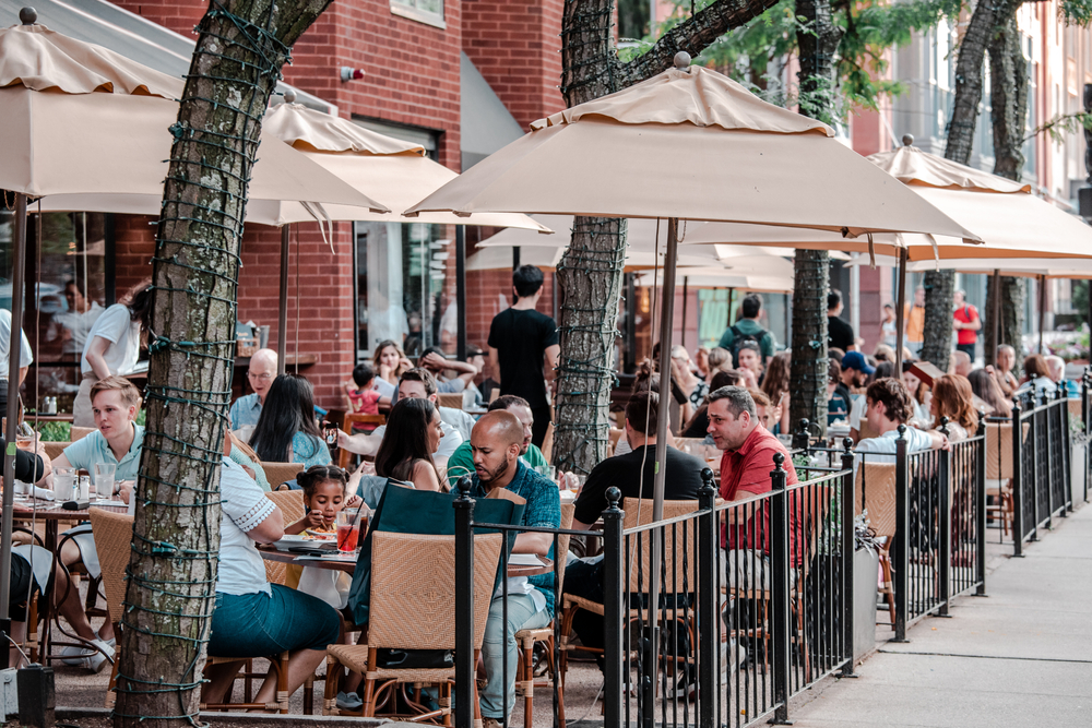A busy restaurant where people are seen eating in an outdoor dining covered with umbrellas, an image for an article about trip cost to Boston.