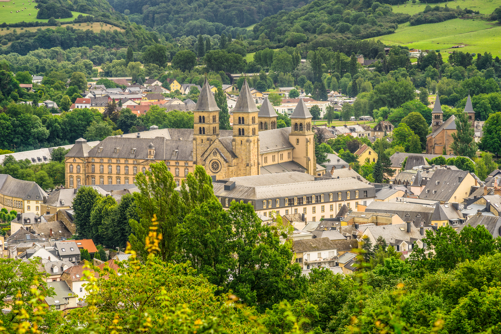 A large old church building in Echternach, a country experience and one of the best areas to stay in Luxembourg, surrounded by small houses, and visible at a distance is a lush forest and vast green field.