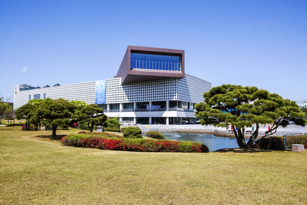 A large building with unique design facing an open field with green grass and a river, a piece on an article about trip cost to South Korea.
