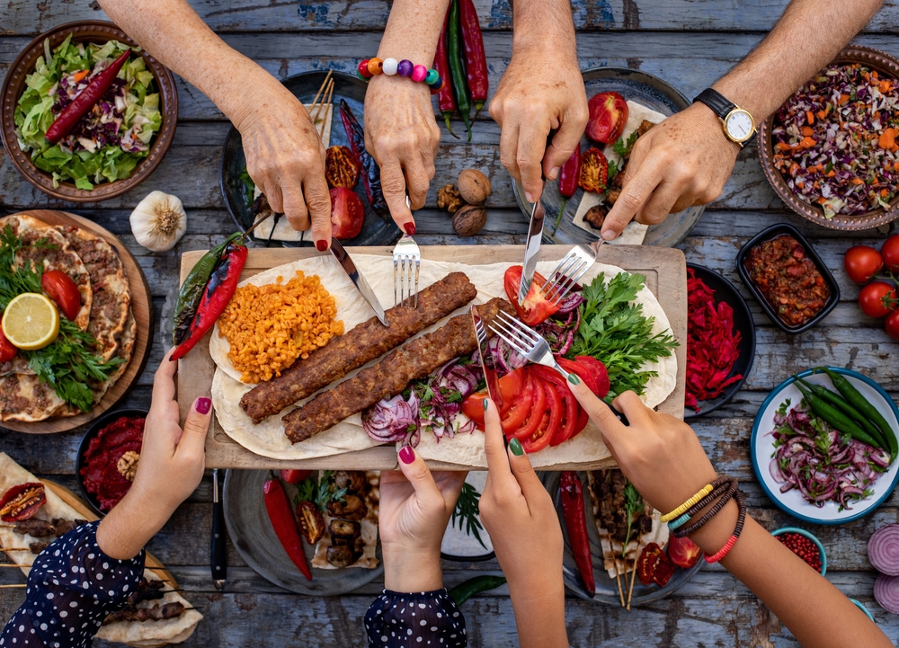 A family sharing a meal where many food can be seen on the table and all of their hands reaching or the food using knife and fork. 