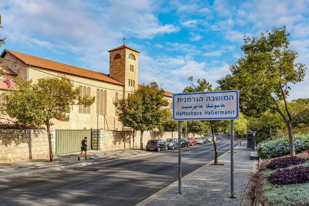 A street sign with a blue Hebrew text and a sidewalk beside an old church with a man jogging and parked cars. 