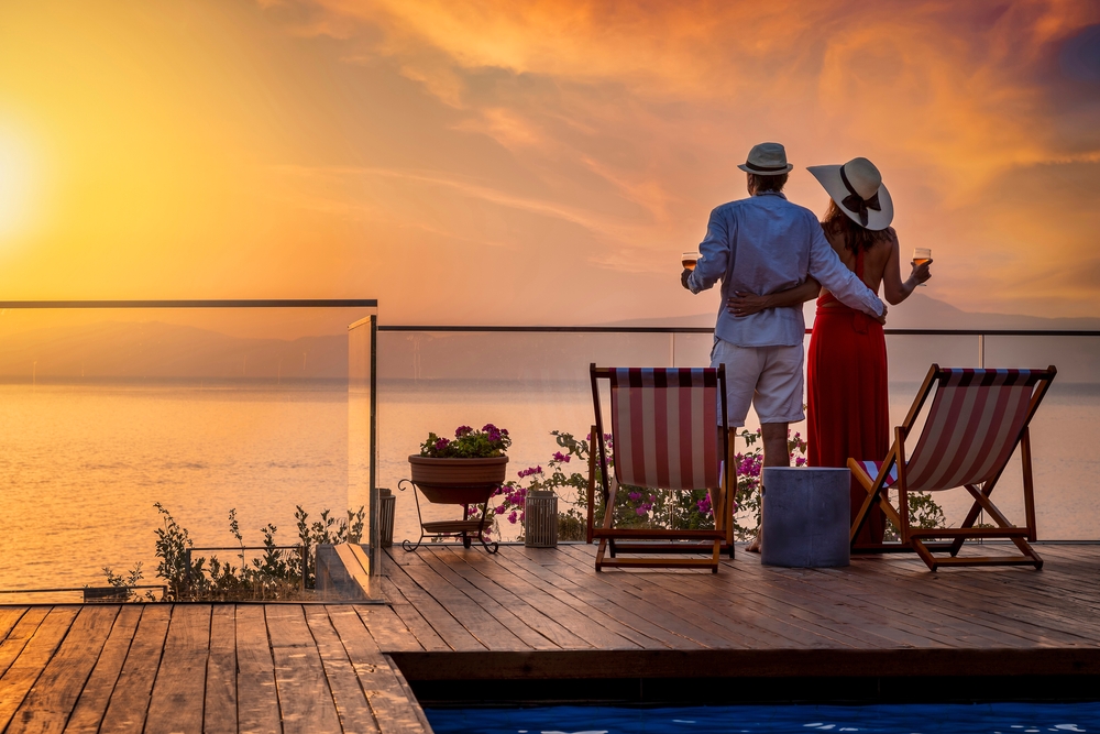 A couple holding each other's waist, holding a glass of wine while staring at the sea during sunset, a photo for where to stay in the Mediterranean.