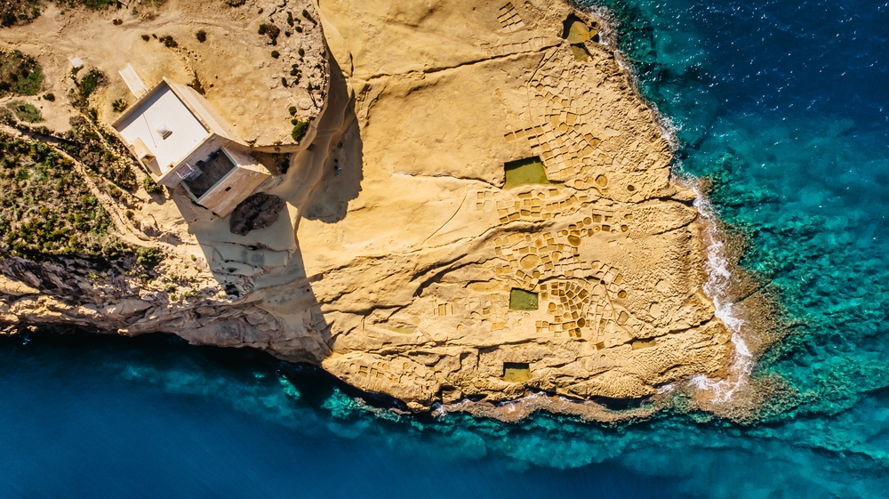Top view of a rocky end of an island in Malta, one of the best places to stay in the Mediterranean, where a lonely solid structure is integrated on the rocks near the clear emerald waters.
