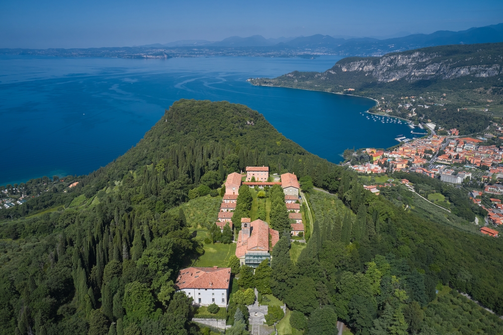 Aerial view of a small town in Bardolino, one of our picks in the best areas to stay in the Italian Lakes, a large portion of the hill beside the lake in covered with a forest and at its peak are several structures, while the plains are populated with residential structures.