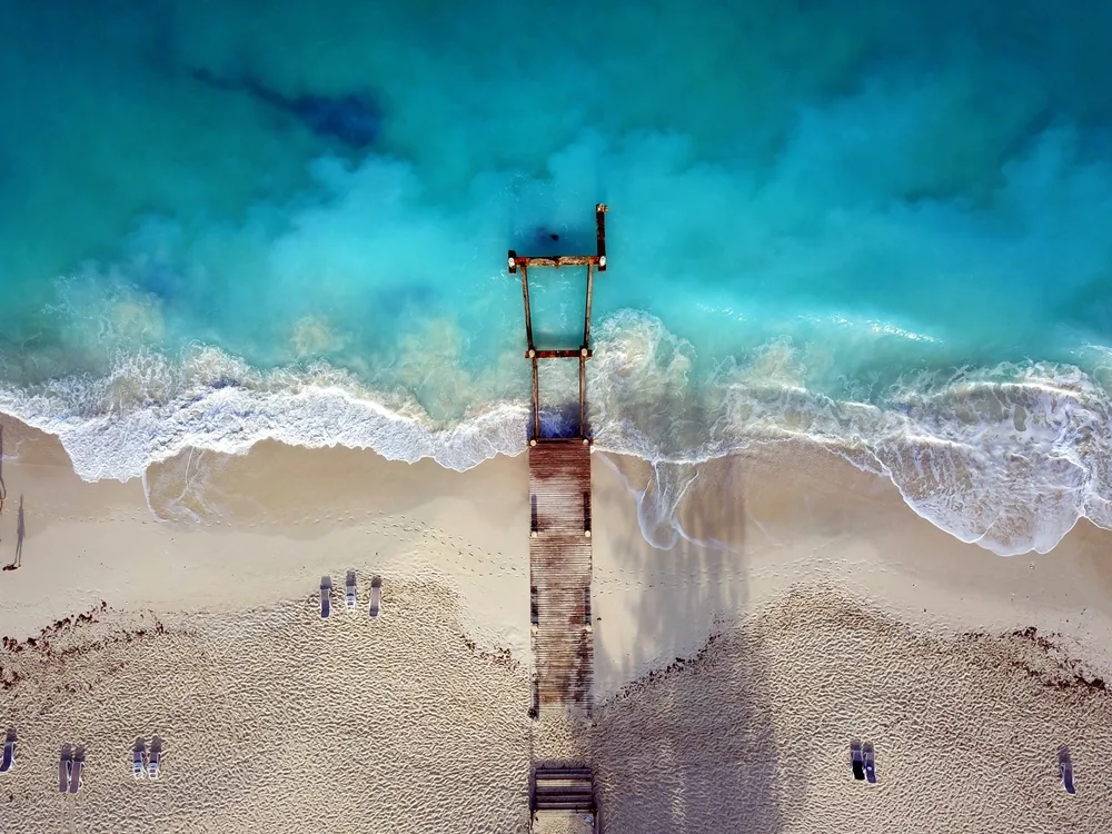 Aerial drone view of a dock on Grace Bay near Club Med with waves washing ashore on Turks and Caicos for a piece answering how long is a flight to Turks and Caicos overall?