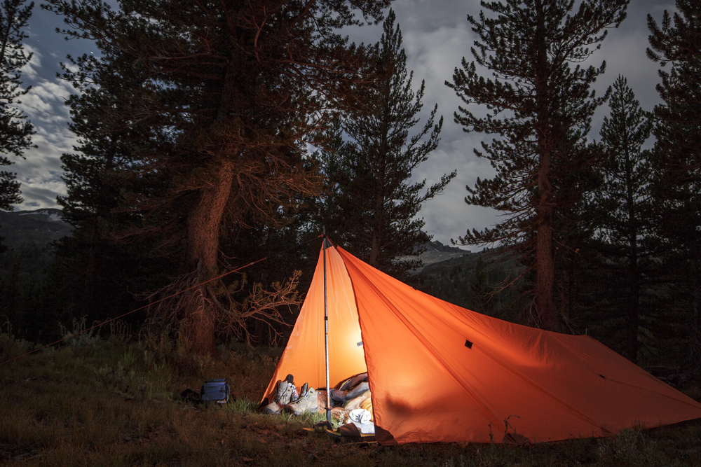 A tent, with a light turned on from the inside, in the middle of a forest during dusk. 