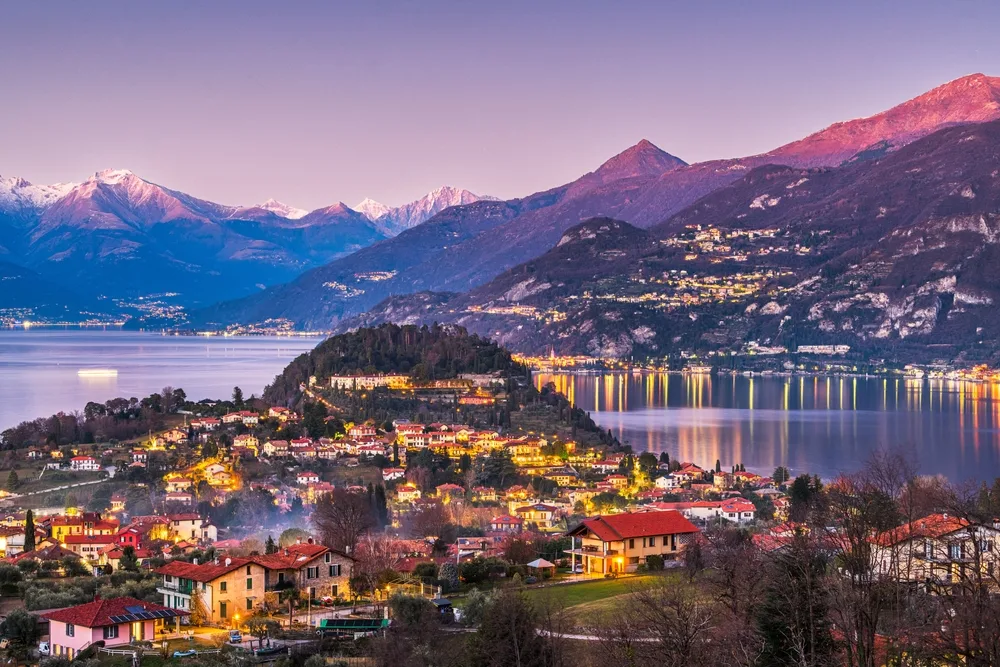 Bellagio, one of the best areas to stay in the Italian Lakes, pictured a town close to the lake at dusk and at a distance are mountains and other residential areas close to the lake.