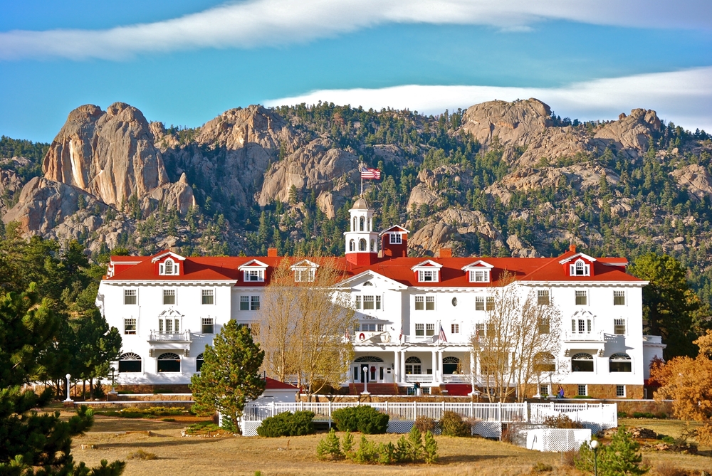 A large white building with red roof and in background are tall mountain with rocks and trees, an image for an article about trip cost to Colorado.