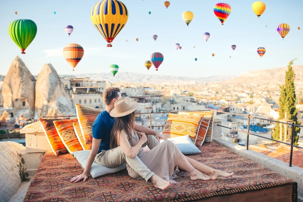 A sweet couple on an outdoor bed staring at the hot-air balloons that are hovering in the air. 