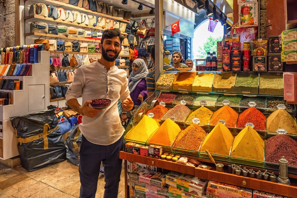 A man posing for a photo in front of his store filled with spices, and the store beside his sells bags in the spice bazaar of Istanbul