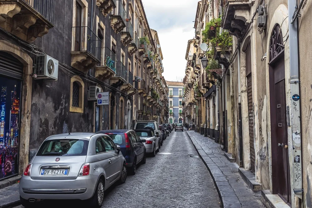 Cars parked in an alley in between 2 old buildings, an image for a travel guide about safety in visiting Sicily.