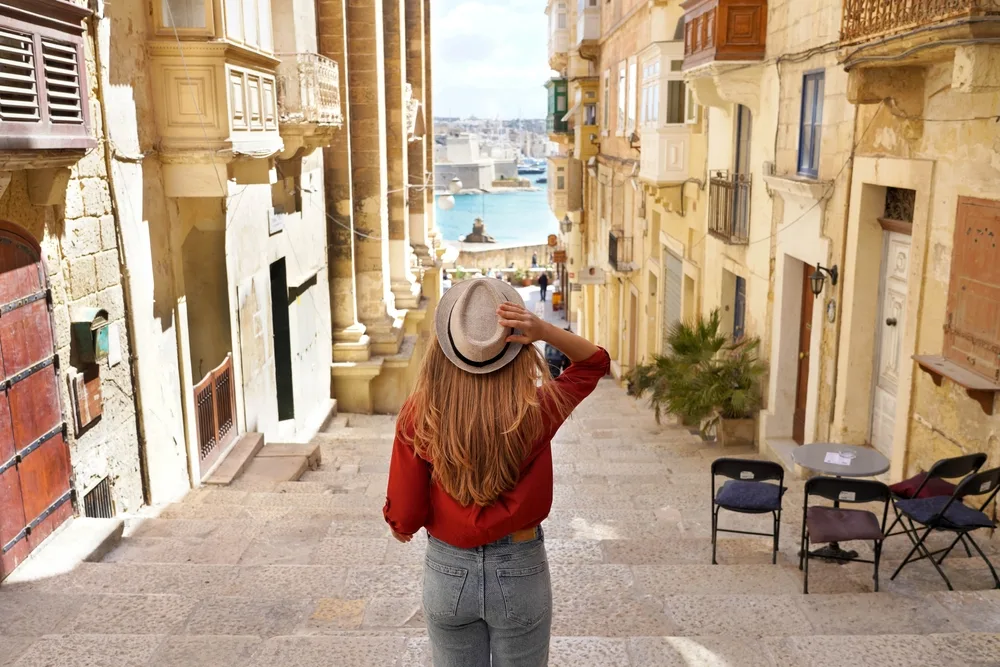 A women wearing red blouse holding her hat over her head while looking ahead a small alley with old building on both sides, photographed for a piece on where to stay in Malta.