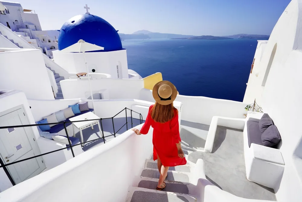 A woman wearing red dress and a hat, walking down the stairs with a view of the vast sea. 