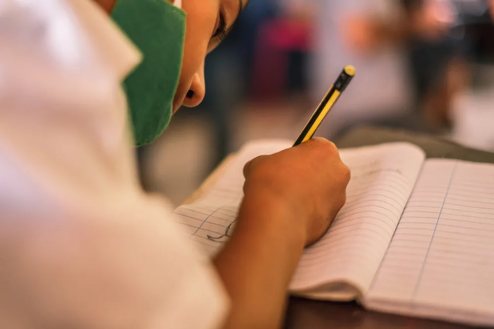 A boy writing on his notebook using a pencil while wearing mask.