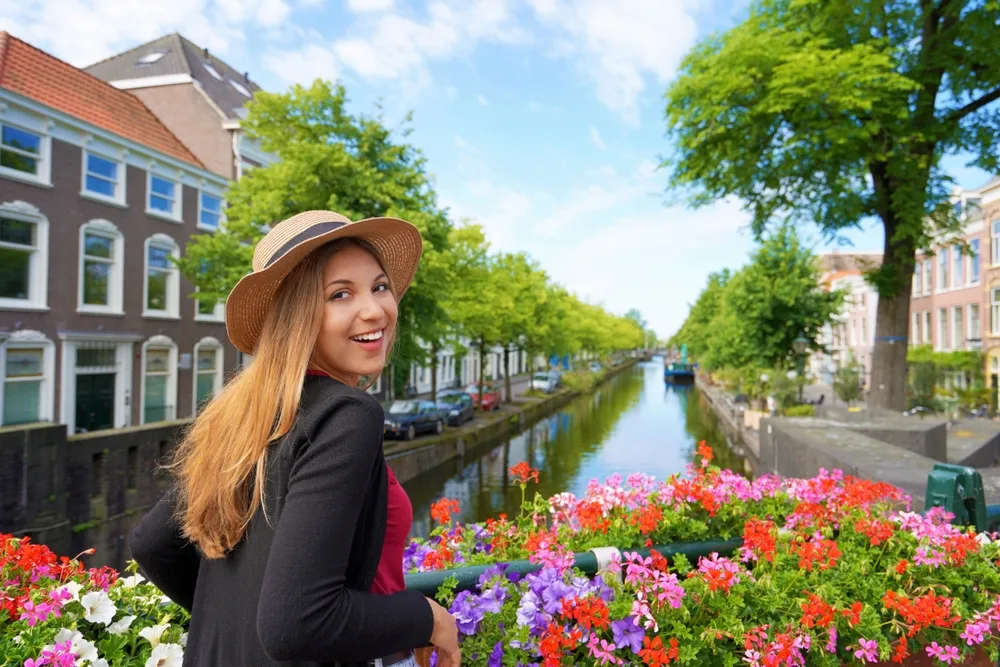 A tourist smiling for a photo a standing on the side of a footbridge decorated with flowers with a view of a long canal, an image for the guide about the safety in visiting the Netherlands.