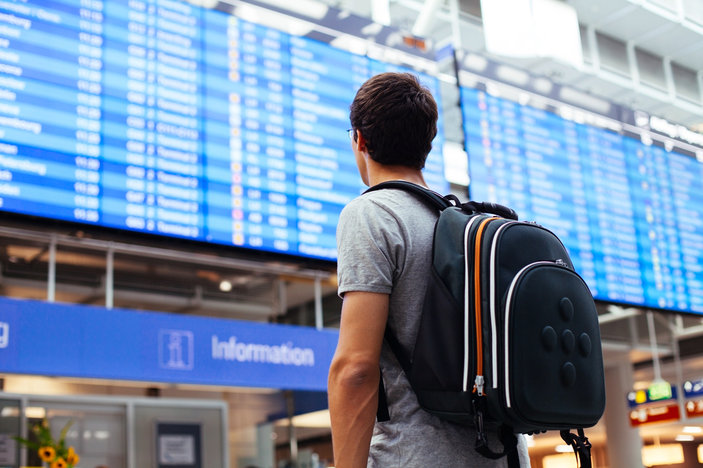 A man wearing his backpack while looking a flight schedules on the screen blurred in background, a concept image for a travel guide about trip cost to Mykonos.