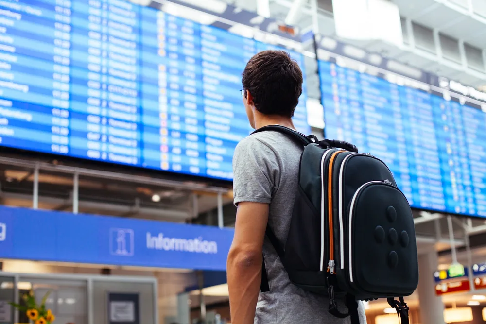 A man wearing his backpack while looking a flight schedules on the screen blurred in background. 