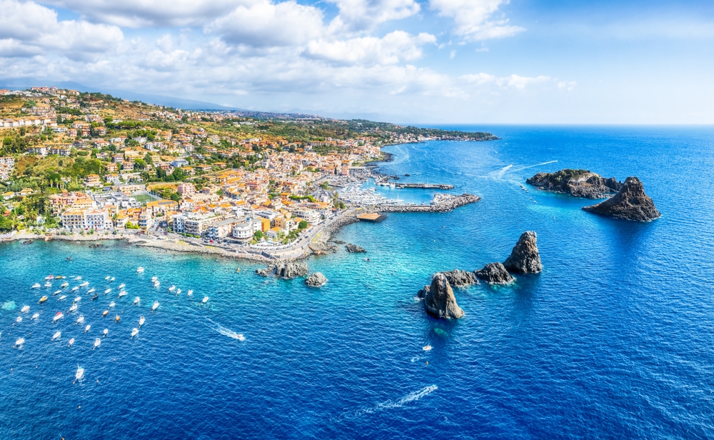 A coastal town in Sicily, Italy, one of the best areas to stay in the Mediterranean,  where hotels are close to the coast, and several boats seen leaving the pier beside large rocks in the middle of the sea. 