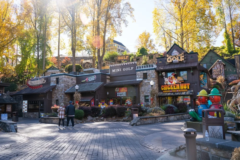 A street with with a view of a restaurant, sports store, and a coffee shop, and a couple walking towards the establishments, an image for a travel guide about trip cost to Gatlinburg.