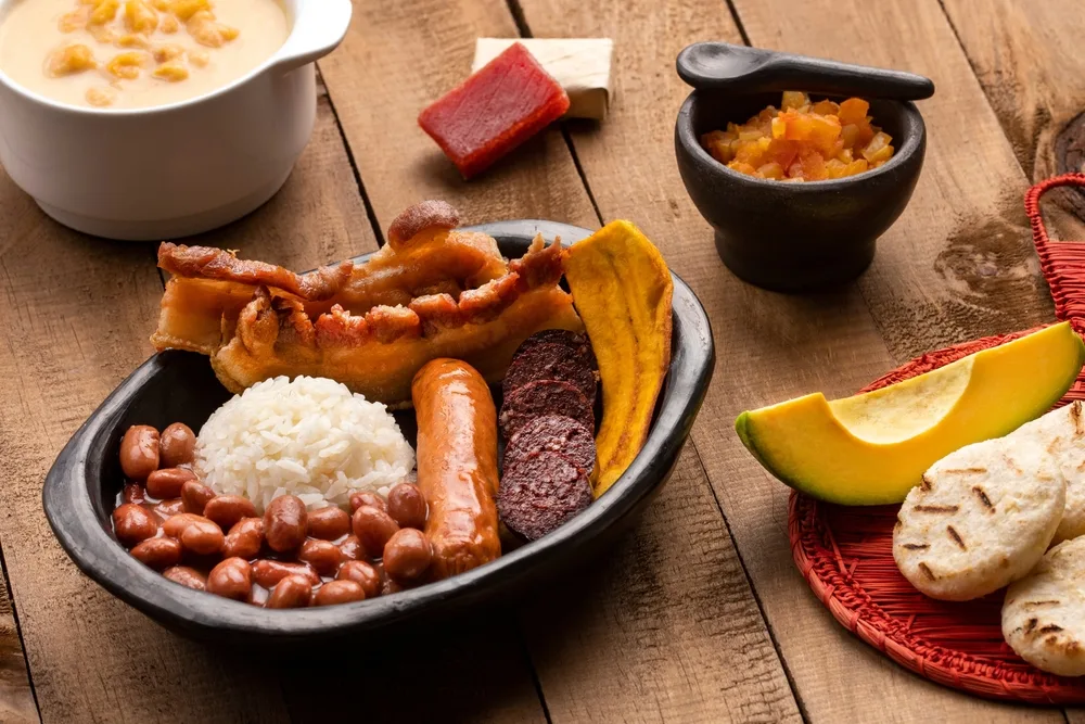 A bowl of meal with rice, beans, sausage, and bacon together with bread, fruits and soup prepared on a wooden table. 
