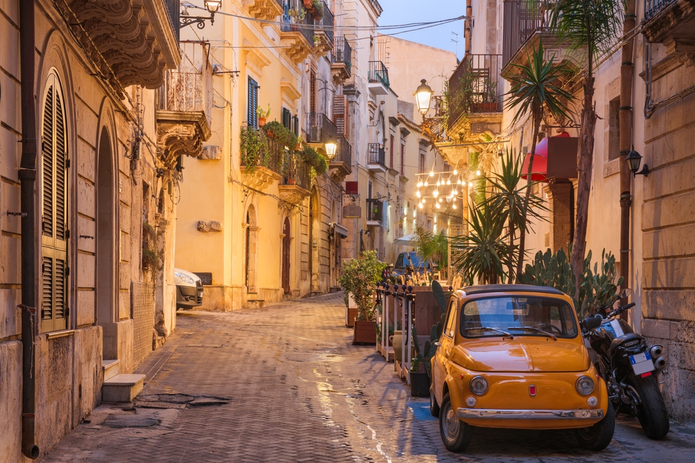 A small alley in between old buildings with a parked small yellow car and a motorcycle, an image for a travel guide about safety in visiting Sicily.