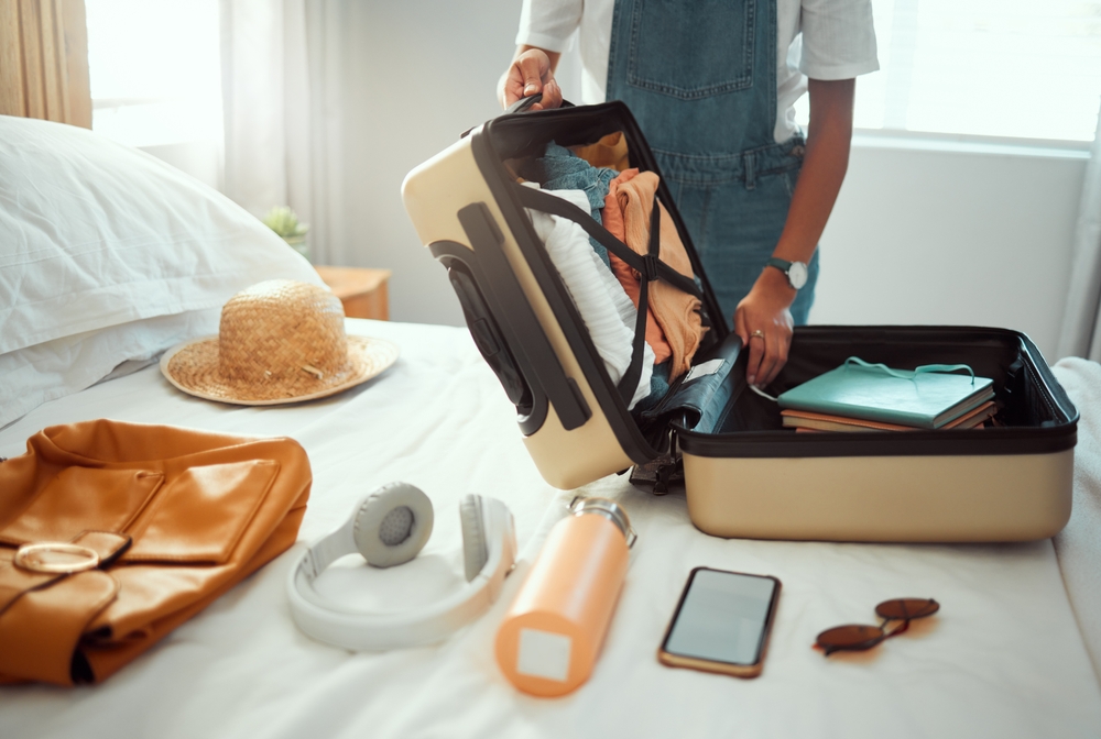 A person can be seen packing her clothes and notebooks inside a luggage where a hat, another bag, wireless headphones, tumbler, phone, and sunglasses can be seen on the bed. 