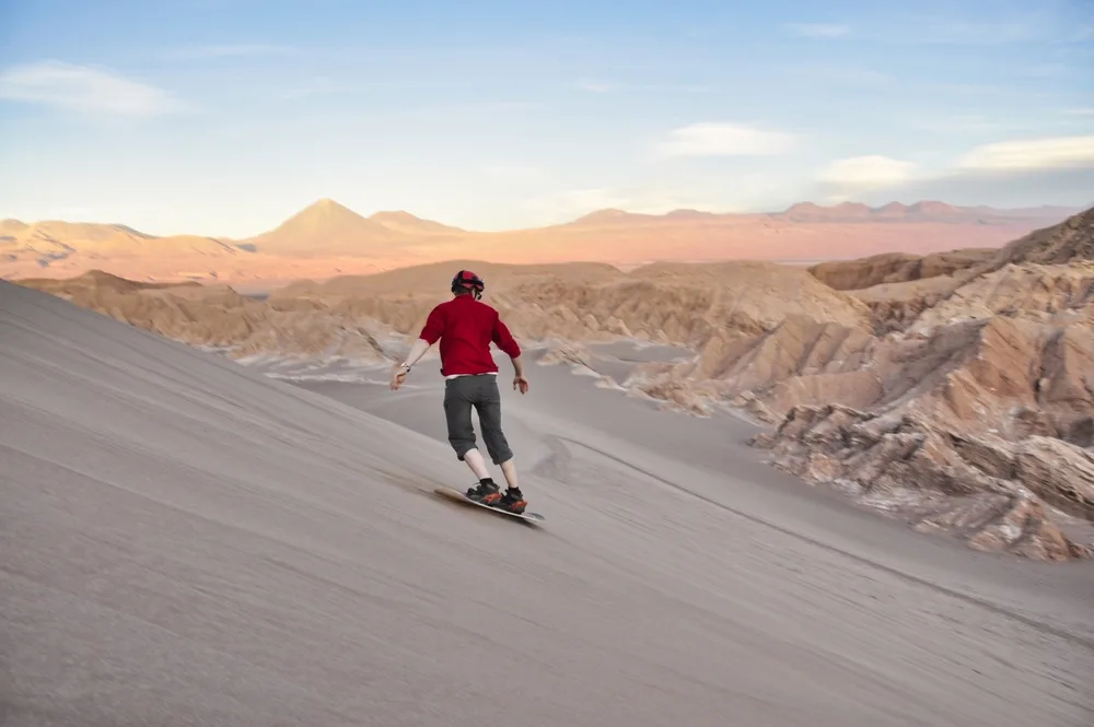 A man wearing red smoothly glides on desert sands while sand boarding, an image for an article about trip cost to Chile.