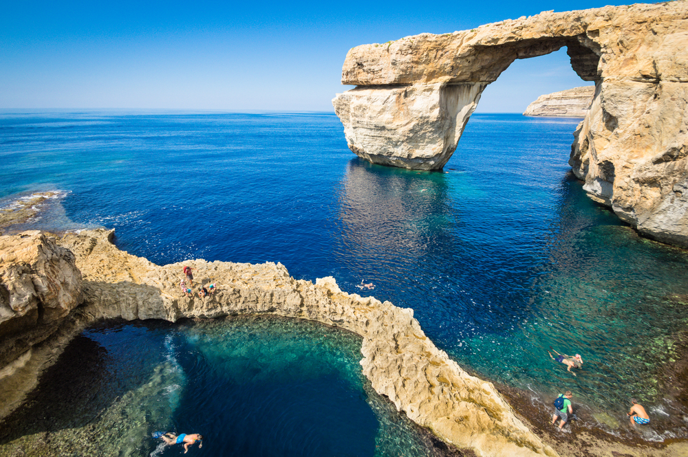 a rock formation on the coastal area in Gozo, one of the best areas to stay in Malta, the rock forms like a bridge and several tourist are snorkelling on the clear waters beside the rock formation.