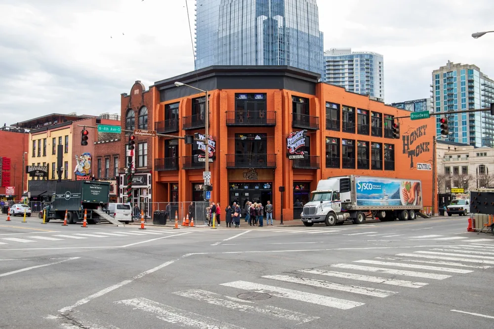 An intersection where a bright orange building, a truck passing by its side and a tall modern building in background, captured for a piece on trip cost to Nashville.