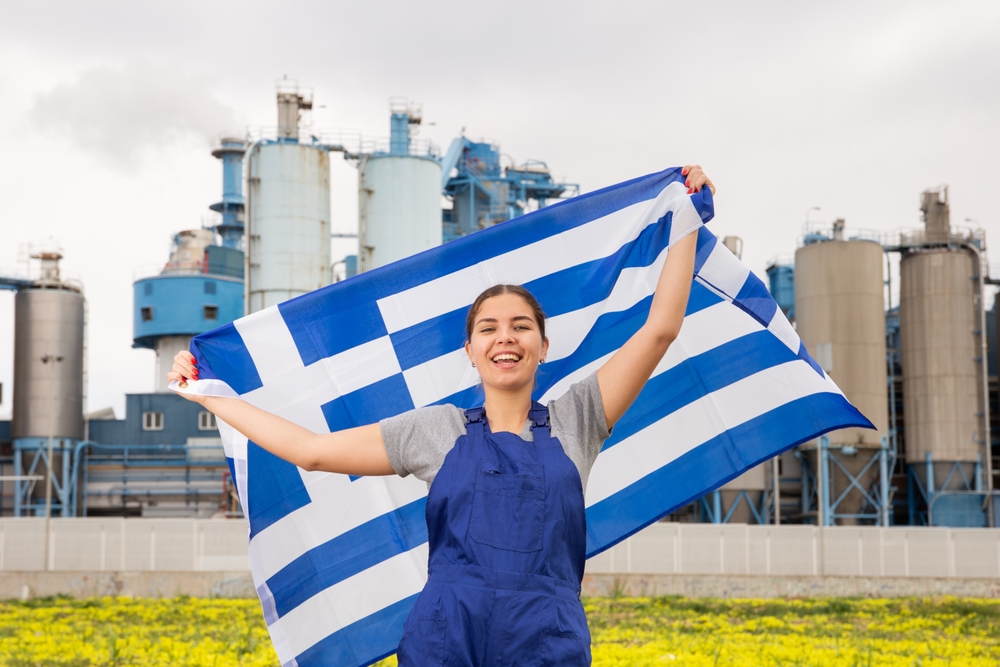 A woman factory worker raising a Greek flag while the factory is behind her, Greece companies are know to be very generous on vacation programs, an item on the list of facts about Greece.