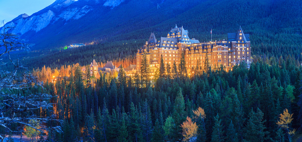 A large hotel structure surrounded by Pine trees with lights turned on during dusk. 