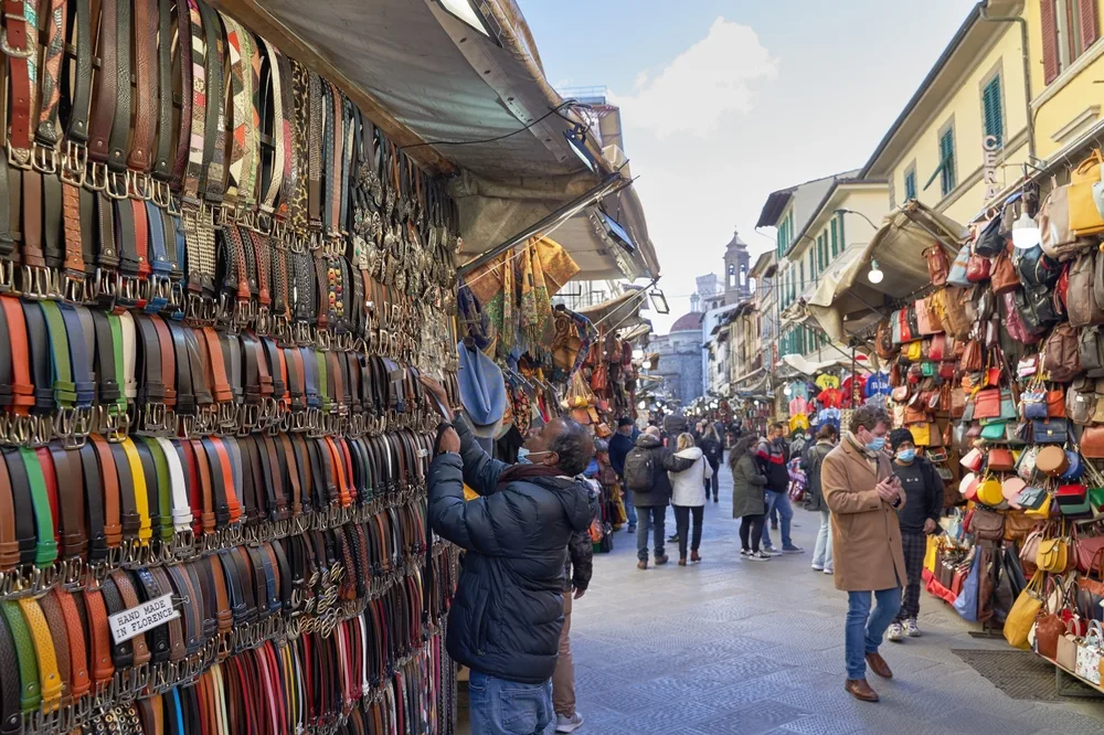 An alley filled with small store stalls selling items such as belts, bags, and other merchandise, an image for the article safety in visit Tuscany.