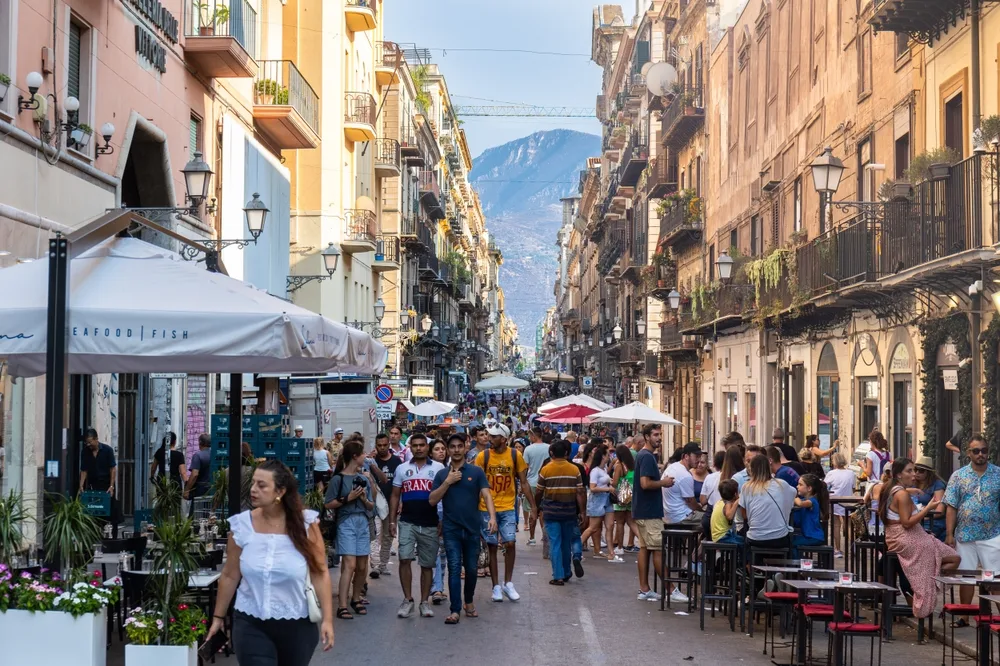 A busy street in between a series of old buildings where some people are walking and others eating in an outdoor dining of restaurants, a section image for a travel guide about safety in visiting Sicily.