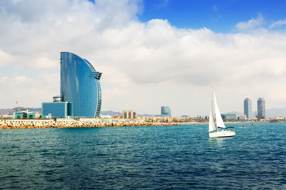 A view from the sea in Barcelona, Spain, one of picks in the best areas to stay in the Mediterranean, where a sailboat and a modern building can be seen during a cloudy afternoon.