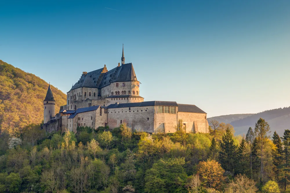 A large old castle on top of a hill surrounded by trees during sunset in Vianden, one of the best areas to stay in Luxembourg.