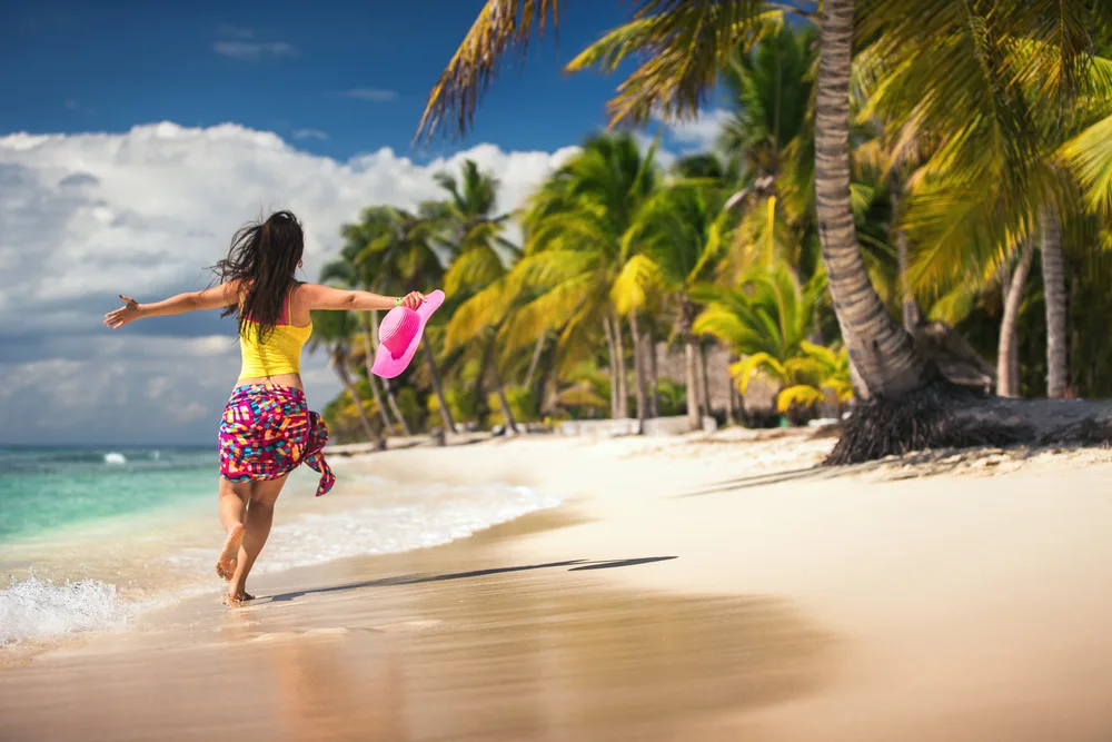 A carefree woman running on the beach with spread arms and holding her pink hat. 