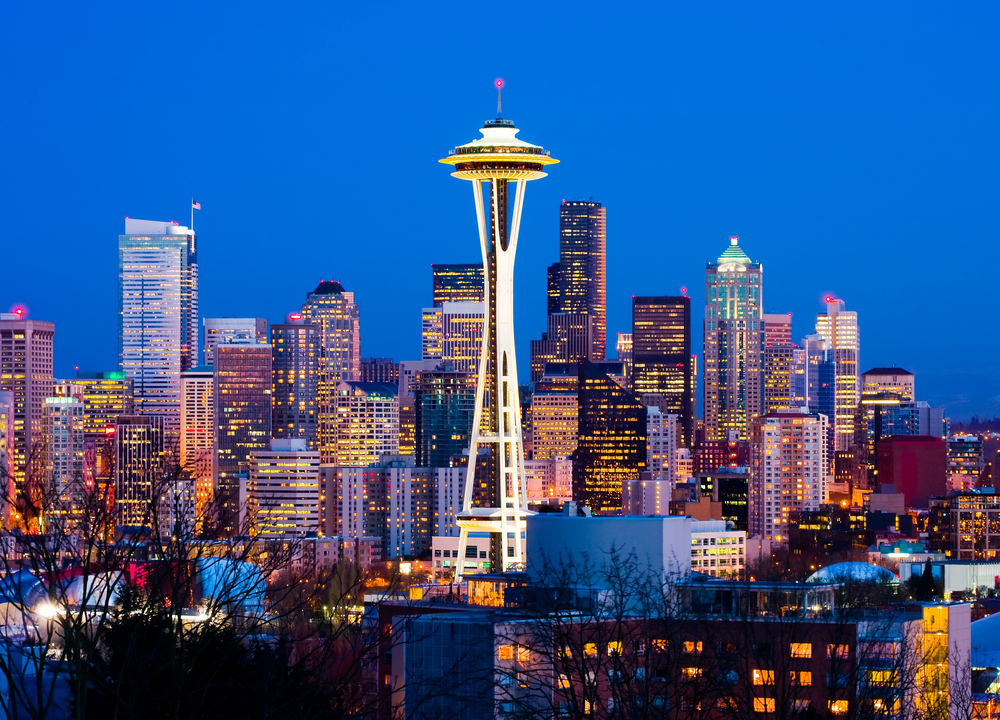 A city skyline during dusk, a piece for an article about trip cost to Seattle, a tall tower stand at the center with a round structure at the top.