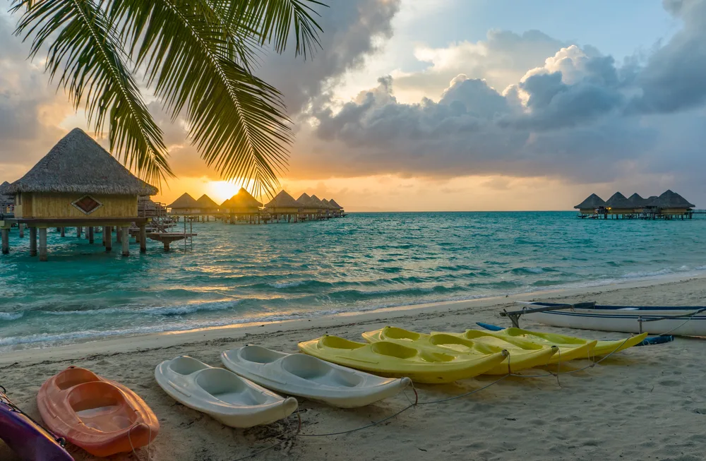 Close-up of a beach on Bora Bora at sunset lined with ocean kayaks with overwater bungalows in the water for an article asking how long is a flight to Bora Bora?