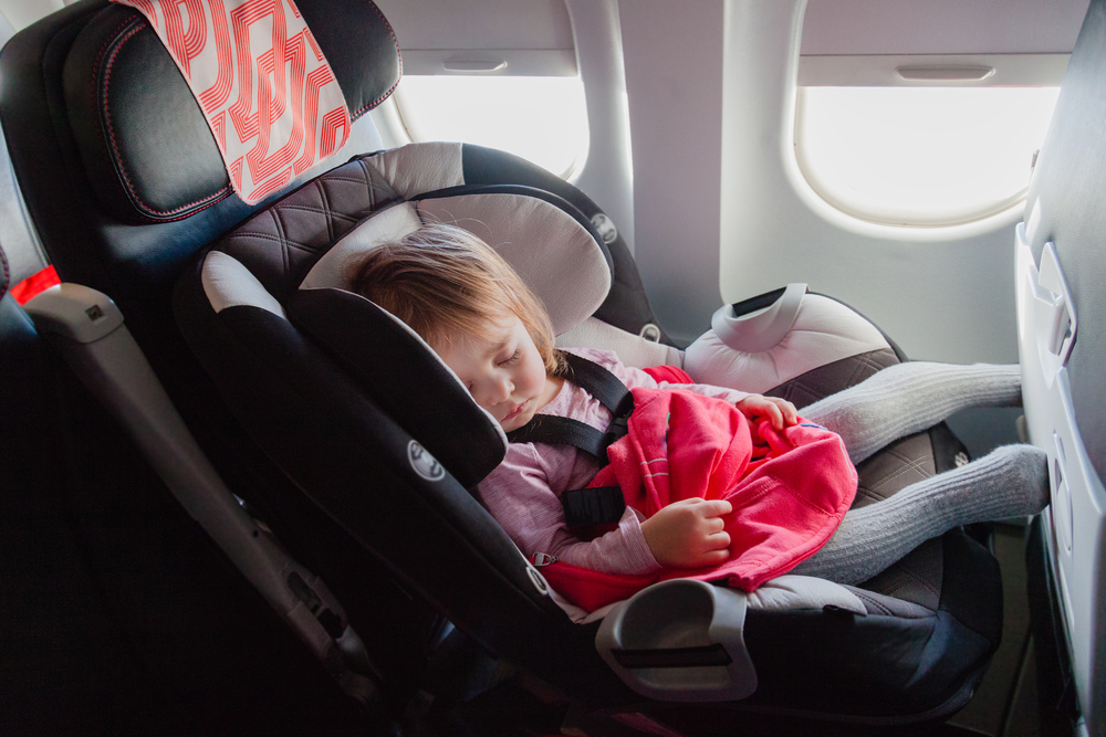 A toddler peacefully sleeping in a car seat placed on an airplane seat, an image for a travel guide about how to travel with a car seat.