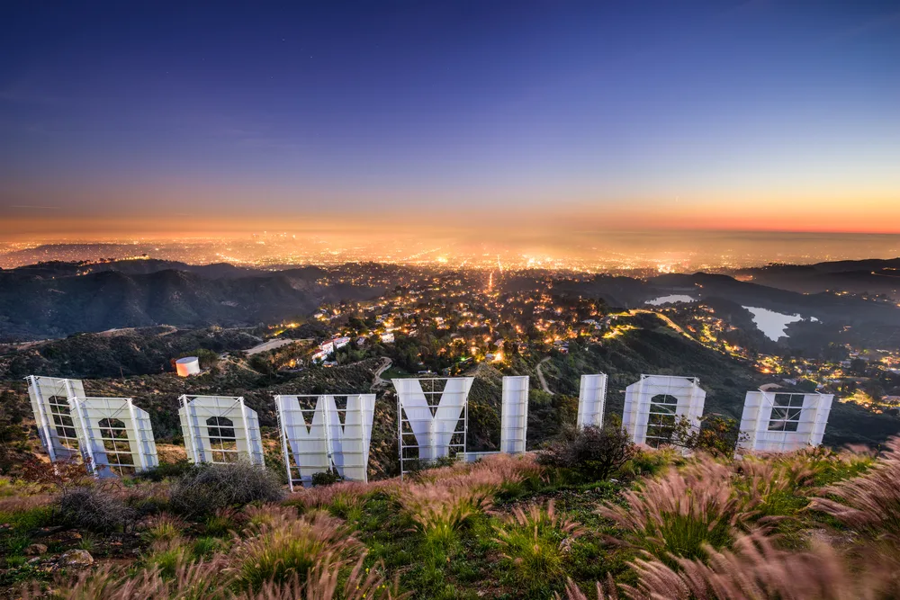 A view behind the famous Hollywood sign of a large city illuminated by lights during dusk. 