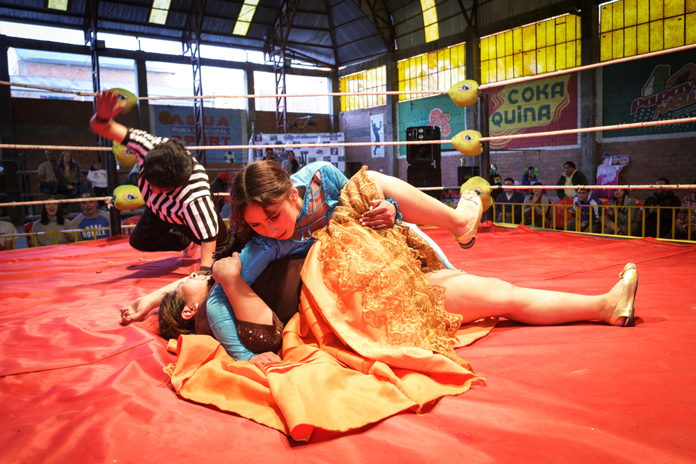 Two women wresting while wearing dress with red-covered ring, and the referee can be seen tapping on the ground. 