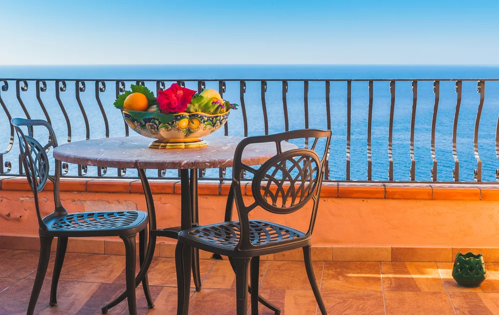 A balcony with small table with a bowl of fruits and two chairs, and a view of the ocean, an image for the article about trip to Sicily cost.