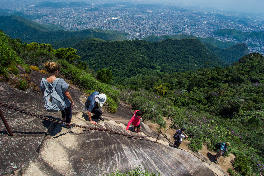 Several people climbing a very steep slope through stair steps carved out from rocks, with the view of a thick forest and a populated city from below.