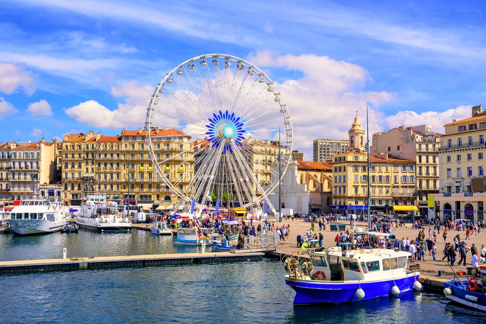 A pier with a dock and several small boats in Marseille, one of the best areas to stay in France, where a lot of people can be seen walking and a Ferris wheel can be seen with low-rise structures in background. 