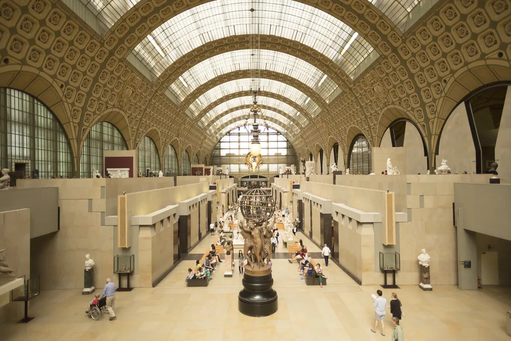 Photograph inside a museum with statue at the center and people can be see seating on benches and some are walking.