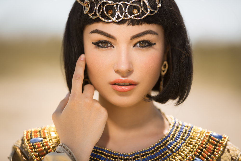 A woman wearing accessorised clothes pointing at her eyes with heavy makeup.