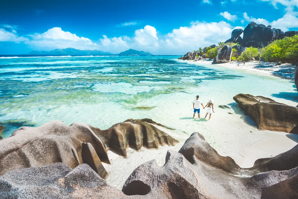 A sweet couple holding hands while walking on a tropical secluded beach with huge rocks on La Digue, our pick on the best areas to stay in Seychelles.