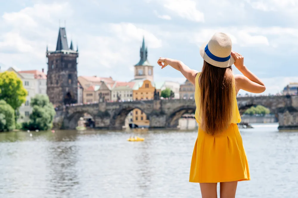 A woman wearing a short yellow dress and a hat points to an old bridge while on the other side of the river. 